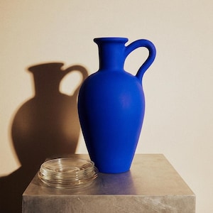 Op de afbeelding: Een kobaltblauwe keramische kruik met een gebogen handvat en een kleine, ronde opening. Een stapel heldere glazen onderzetters staat voor de kruik op een zilveren metalen kubus. De kruik werpt een schaduw op de beige muur.