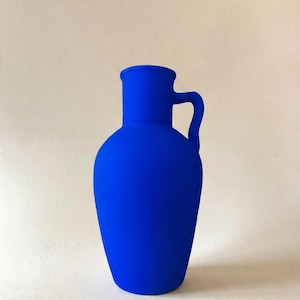 May include: A vibrant cobalt blue vase with a matte finish, featuring a rounded body, a narrow neck with a flared rim, and a single curved handle attached to the side. The vessel has a solid, opaque appearance with a smooth, uniform texture.