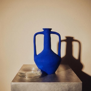 May include: A cobalt blue vase with two handles and a stack of clear glass plates on a metallic surface. The vase casts a shadow on the beige wall behind it. The vase has a matte finish.
