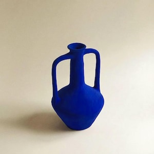 Puede incluir: Un jarrón de cerámica azul cobalto con dos asas y un cuello estrecho. El jarrón tiene un cuerpo redondeado y un acabado mate. El diseño recuerda a las ánforas griegas antiguas. Un artículo de decoración para el hogar.