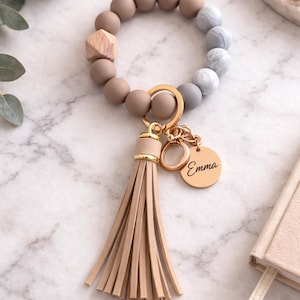 May include: A beaded wristlet keychain with a wooden accent and a tassel. The wristlet features a mix of taupe, gray, and marble-patterned beads. A gold-colored charm with the name "Emma" is attached to the keychain.