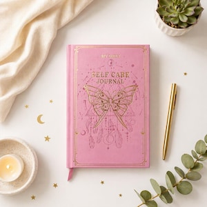 May include: A pink "Self Care Journal" with gold foil details, featuring a butterfly design and the text "My Daily." A gold pen, a small succulent, and a lit candle are also visible.