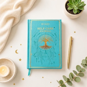May include: A teal-colored journal with gold accents and the words "My Daily Self Care Journal" on the cover. The cover features a gold tree design and a dreamcatcher motif. A gold pen rests beside the journal, with a succulent and candle in the background.