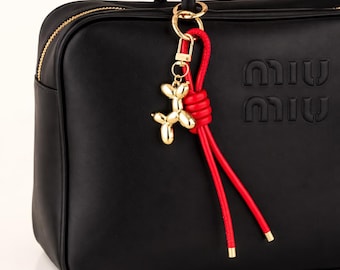 May include: A black leather handbag featuring a gold-tone zipper and the embossed text MIU MIU. Attached to the handle is a gold-tone metal balloon dog charm and a red leather cord knotted into a loop with two dangling ends and gold-tone tips.
