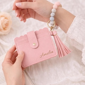 May include: A light pink cardholder with a scalloped edge, snap closure, and the name "Amelia" in gold script. It is paired with a matching tassel keychain and a beaded wristlet in pastel colors.