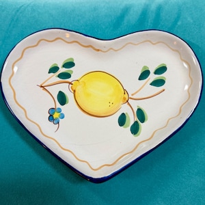 Puede incluir: Un plato de cerámica en forma de corazón con base blanca y borde azul oscuro. El centro presenta un limón amarillo pintado a mano con hojas verdes y una pequeña flor azul sobre un tallo marrón. Una línea ondulada de color marrón claro sigue el borde.