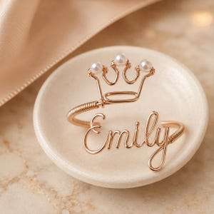 May include: A gold-toned wire cuff bracelet featuring the name Emily in cursive script and a crown motif adorned with three small white pearls. The bracelet has a coiled wire design on the sides and an open back for adjustable wear.