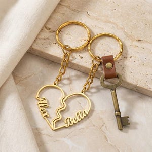 Puede incluir: Llaveros dorados con un colgante en forma de corazón dividido en dos, cada mitad con un nombre, y una llave vintage con una correa de cuero marrón. Los llaveros están conectados por cadenas y anillos dorados.