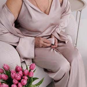 May include: A light mauve satin pajama set, including a camisole, pants, and a long-sleeved robe. The set is worn by a person sitting in a white chair. A bouquet of pink tulips is in the foreground.