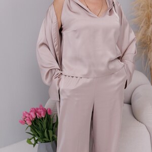 May include: A light mauve satin pajama set. The set includes a hooded top with a V-neck and a matching pair of wide-leg pants. The top has long sleeves and a relaxed fit. The fabric has a subtle sheen. The outfit is suitable for sleepwear or loungewear.