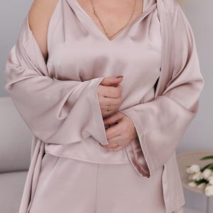 May include: A light pink satin pajama set, including a camisole, shorts, and a long-sleeved robe. The set features a V-neck camisole and wide sleeves on the robe. A gold necklace and rings are visible.