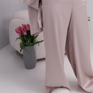 May include: A light mauve-colored pajama set, featuring wide-leg pants and a matching top. A gray vase holds pink tulips, and a white chair is in the background. The outfit is made of a soft, flowing fabric.