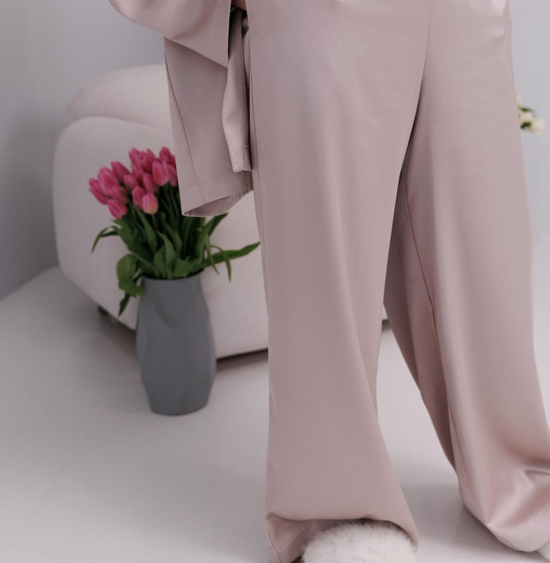 May include: A light mauve-colored pajama set, featuring wide-leg pants and a matching top. A gray vase holds pink tulips, and a white chair is in the background. The outfit is made of a soft, flowing fabric.