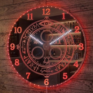 May include: A circular wall clock with a mirrored surface and red LED illumination. The clock face features red Arabic numerals from 1 to 12 and a central circular design with etched symbols and runic-style characters. Two white hands indicate the time.