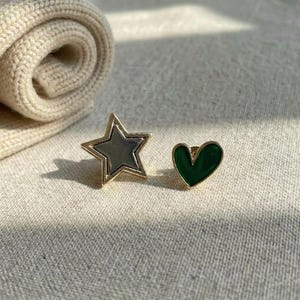 Könnte beinhalten: Ein Paar Ohrringe, einer in Sternform und einer in Herzform. Der Stern hat einen goldfarbenen Umriss und eine silberfarbene Mitte. Der Herzohrring ist grün mit einem goldenen Umriss. Die Ohrringe werden auf einer strukturierten, hellen Oberfläche präsentiert.