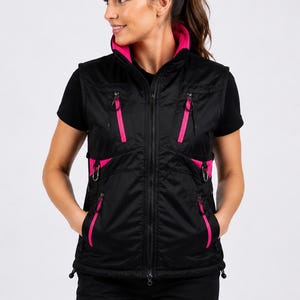 May include: A black sleeveless vest featuring a high collar with a bright pink interior lining. The garment includes multiple zippered pockets with pink trim, a central front zipper, and adjustable drawstrings at the hem for a secure fit.