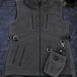 May include: A dark gray sleeveless fleece vest with a high collar and a full-length front zipper. The vest features multiple zippered pockets, black webbing straps with metal D-rings on the sides, and a detachable small pouch with a metal clip.