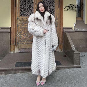 May include: A long, luxurious white fur coat with a chevron pattern. The coat has a large collar and wide sleeves. The model is wearing pink bow-adorned heels and holding a silver clutch. The coat is floor-length.