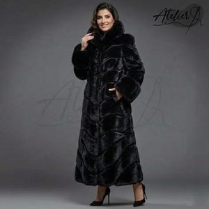 Puede incluir: Abrigo largo de piel sintética negra con capucha y mangas largas. El abrigo presenta un patrón texturizado en forma de ondas y se abrocha con botones. La modelo lleva tacones altos negros. El logo de Atelier J está en la esquina superior derecha.