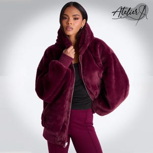 May include: A burgundy faux fur bomber jacket with a high collar and a silver zip. The jacket has long sleeves and a ribbed hem. The Atelier J logo is in the upper right corner. The model is wearing a black top and burgundy trousers.