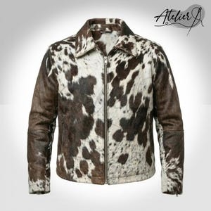 May include: A full-zip jacket with a cowhide pattern in shades of brown and white. The jacket has a collared neckline and two side pockets. The brand name "Atelier J" is written in cursive on the upper right corner.