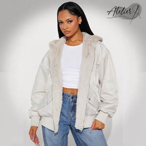 May include: A cream-colored bomber jacket with a faux fur-lined hood and collar. The jacket has a quilted design, ribbed cuffs, and a zipper closure. The model is wearing blue jeans and a white top. The Atelier J logo is in the upper right corner.