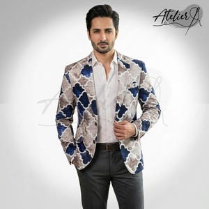 May include: A blazer featuring a blue and beige quatrefoil pattern, worn over a crisp white button-down shirt and dark gray trousers. The garment includes a notched lapel and a chest pocket. The text Atelier J is visible in the upper right corner.