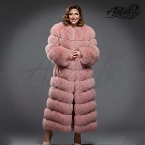 May include: A full-length, light pink fur coat with a round collar. The coat has a horizontal design and is worn by a person. The coat is floor-length and has a soft, luxurious appearance. The image includes the text "Atelier J" in the upper right corner.