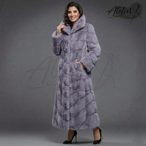 May include: A full-length, light purple fur coat with a high collar and long sleeves. The coat has a textured pattern and a button-up front. The woman in the image is wearing black high heels. The coat is from Atelier J.