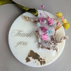 May include: A circular, cream-coloured decorative piece inscribed with "Thank you" in cursive. It features dried flowers in pink, yellow, and blue, and a green ribbon. Gold leaf details add a luxurious touch to the design.