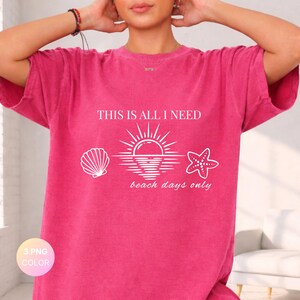 Può includere: T-shirt magenta con la scritta "THIS IS ALL I NEED" e la frase "beach days only". Il design include una conchiglia, un sole sull'acqua e una stella marina. Un capo d'abbigliamento casual.