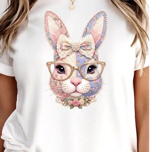 Può includere: T-shirt bianco con un design a coniglio stravagante. Il coniglio presenta un motivo patchwork rosa e blu, adornato con un fiocco, occhiali e un colletto floreale. Il design è dettagliato con pizzo e dettagli floreali, creando un look affascinante e artistico.