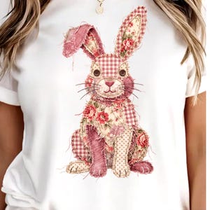 Puede incluir: Camiseta blanca con un diseño de conejo patchwork. El conejo está hecho de varias telas, incluyendo cuadros, flores y lunares, en tonos rosa, rojo y crema. El conejo tiene ojos de botón y bigotes, creando un aspecto artesanal.