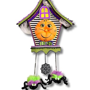 May include: A decorative wall clock shaped like a house with a black and white striped background, a purple roof, and a central orange pumpkin face. It features a green and black striped bow tie, a hanging spider web, and two fuzzy spiders.