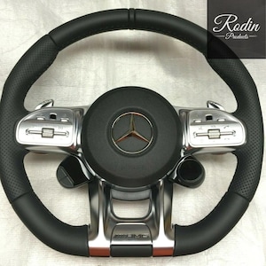 Puede incluir: Volante de cuero negro con detalles plateados y un logotipo de Mercedes-Benz. Cuenta con empuñaduras de cuero perforado y botones de control. El logotipo AMG es visible. Un producto de Rodin Products.