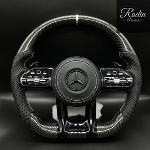 Può includere: Un volante nero e in fibra di carbonio con il logo Mercedes-Benz al centro. Presenta cuciture bianche, dettagli argentati e pulsanti di controllo. Il marchio "Rodin Products" è visibile in alto a destra.