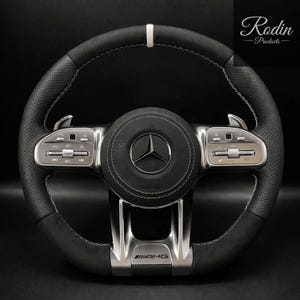 May include: Black and silver Mercedes-Benz AMG steering wheel with white stitching. The wheel features a perforated leather grip, silver control buttons, and the Mercedes-Benz logo. The AMG logo is visible at the bottom.