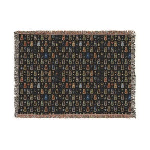 May include: A black woven throw blanket with a repeating pattern of colorful, stylized cat figures and other symbols. The blanket has a fringed edge in shades of brown and tan. The cat figures are in various colors, including red, blue, and green.