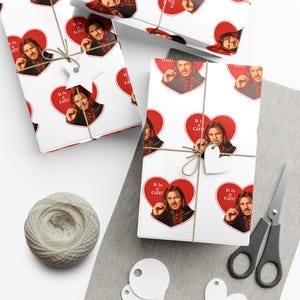 May include: White wrapping paper with a repeating pattern of a man's image inside a red heart with the text "It is a Gift!". The wrapped gifts are tied with twine and have heart-shaped gift tags. Scissors and a ball of twine are also visible.