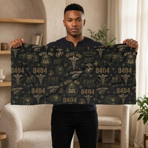 May include: A rectangular black towel featuring a repeating pattern of military-themed graphics in olive green. The design includes the text 8404, Devil Doc, Corpsman Up, and FMF, alongside illustrations of medical bags, caduceus symbols, and bulldogs.
