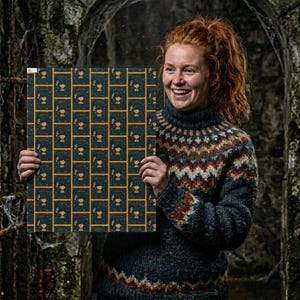 Op de afbeelding: Een vierkante print met een herhalend patroon van kleine illustraties op een donkerblauwe achtergrond met gouden randen. De illustraties tonen een figuur met een boek en een kleine vlam. De print wordt vastgehouden door een persoon die een trui met patroon draagt.