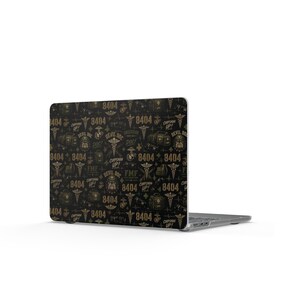 Op de afbeelding: Een laptop met een zwart en goudkleurige patroonsticker met de tekst 8404, Devil Doc en Corpsman Up, samen met medische caduceussymbolen, militaire emblemen en illustraties van medische tassen in een herhalend grafisch ontwerp.