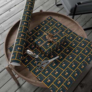 Op de afbeelding: Op een houten tafel liggen cadeaupapier, een ingepakt cadeau en een rol papier. Het donkerblauwe papier heeft een herhalend patroon van een stripfiguur en de naam "CORALINE" in goud. Een schaar en plakband zijn ook aanwezig.