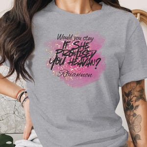 May include: A grey t-shirt featuring the text "Would you stay IF SHE PROMISED YOU HEAVEN? Rhiannon" in black script over a pink and gold glitter design. The sleeves of the shirt are rolled up.