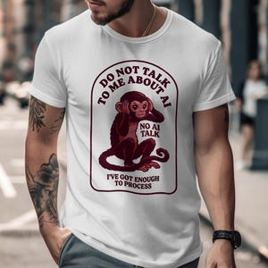 以下が含まれることがあります： 白いTシャツに、耳をふさぐ猿のマルーン色のグラフィック。Tシャツのテキストは「DO NOT TALK TO ME ABOUT AI」と「I'VE GOT ENOUGH TO PROCESS」と書かれています。「NO AI TALK」というフレーズも含まれています。
