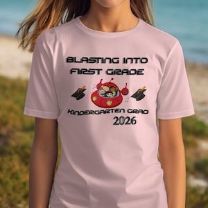 Puede incluir: Camiseta rosa claro con el texto "BLASTING INTO FIRST GRADE" sobre un gráfico de nave espacial roja con niños. Debajo del gráfico está "Kindergarten Grad 2026". La camiseta celebra la graduación de un niño de jardín de infancia.
