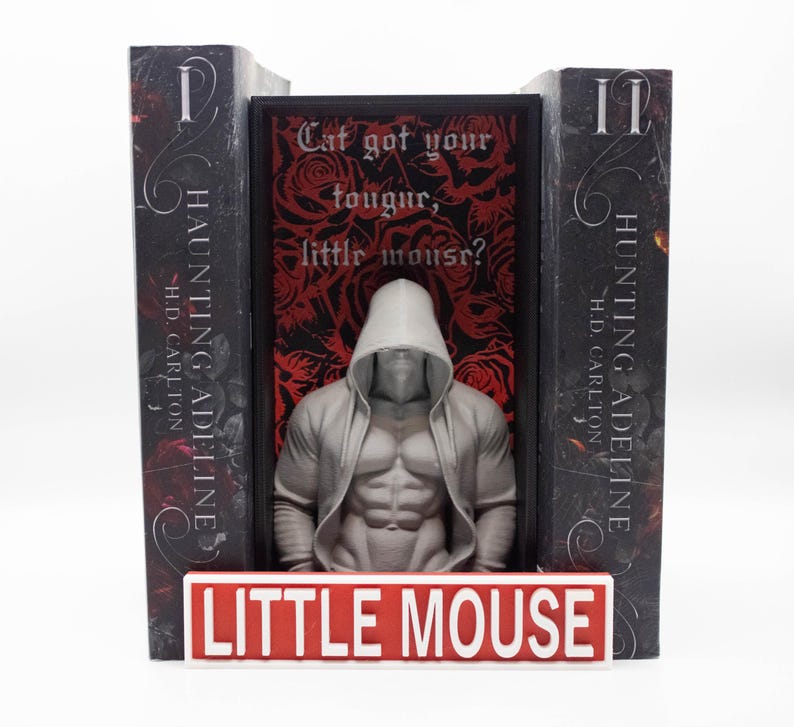 May include: A bookend set featuring two books with the title "Haunting Adeline" and a central figure of a muscular person in a hooded sweatshirt. The base reads "LITTLE MOUSE" in red and white. The background has text that reads "Cat got your tougur, little mouse?"