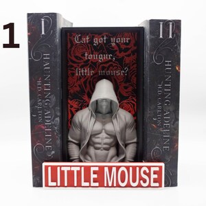 May include: A bookend set featuring two dark gray bookends with the text "HAUNTING ADELINE" and "HUNTING ADELINE". A central figure of a muscular person in a hooded jacket stands between the bookends. The base reads "LITTLE MOUSE".