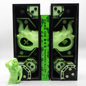May include: A neon green figure of a person in a Scream mask stands next to a book with the title "Game On" and two black bookends. The bookends have a neon green design with a monster face and geometric shapes. The book is by Navessa Allen.