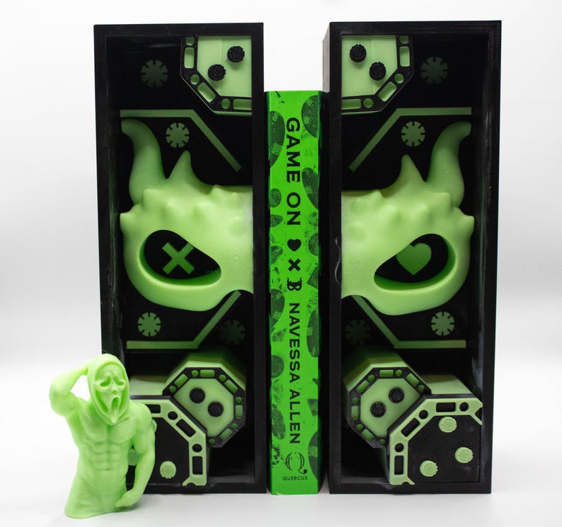 May include: A neon green figure of a person in a Scream mask stands next to a book with the title "Game On" and two black bookends. The bookends have a neon green design with a monster face and geometric shapes. The book is by Navessa Allen.